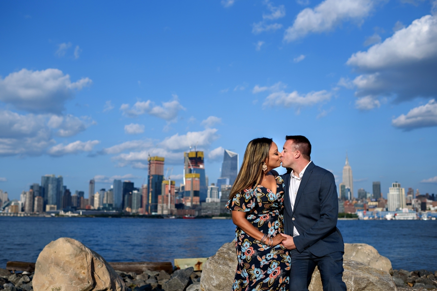 A couple sit on the rocks and kiss with NYC Skyline behind them.